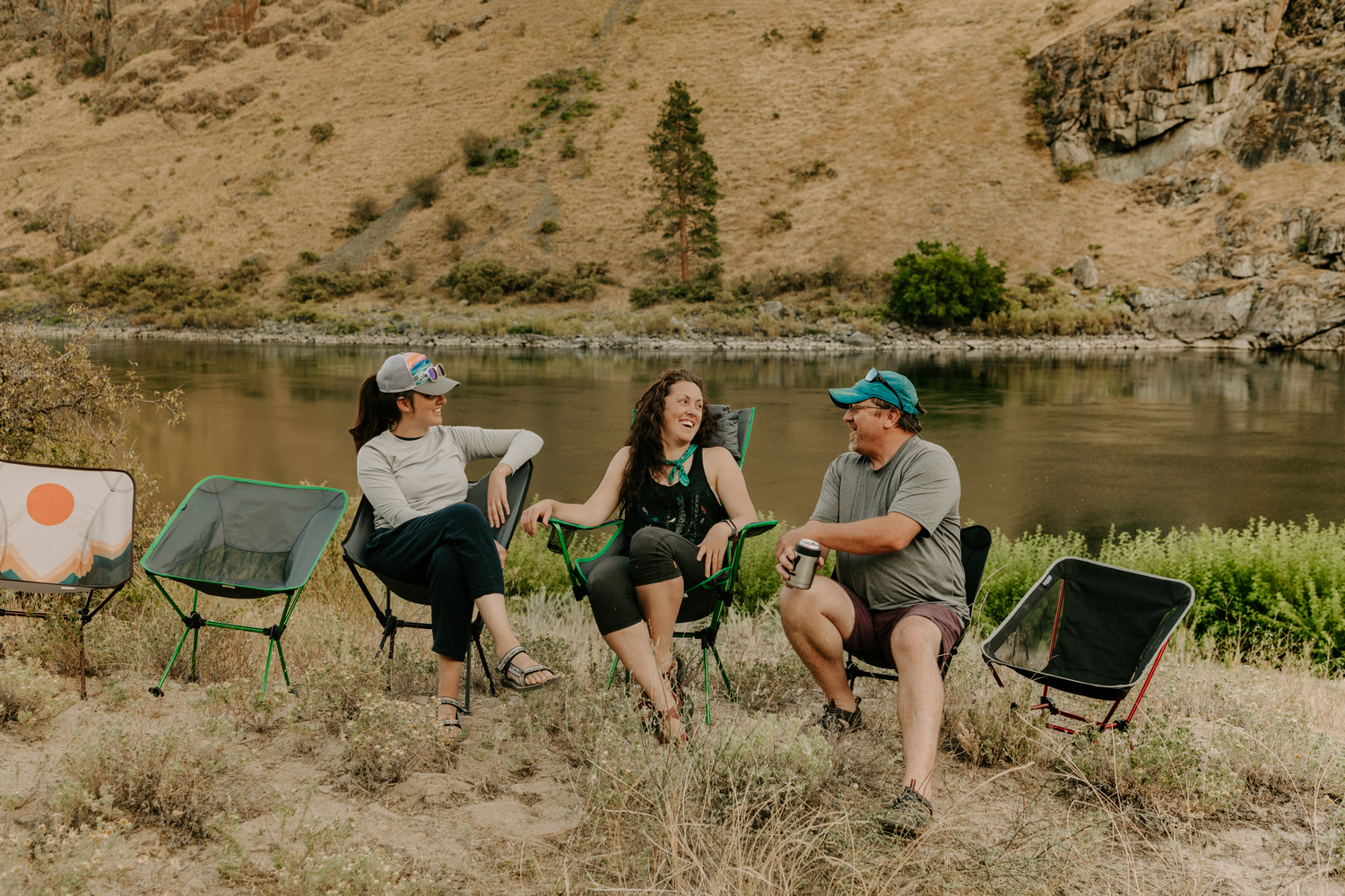 three people outdoors in chairs laughing with river behind them