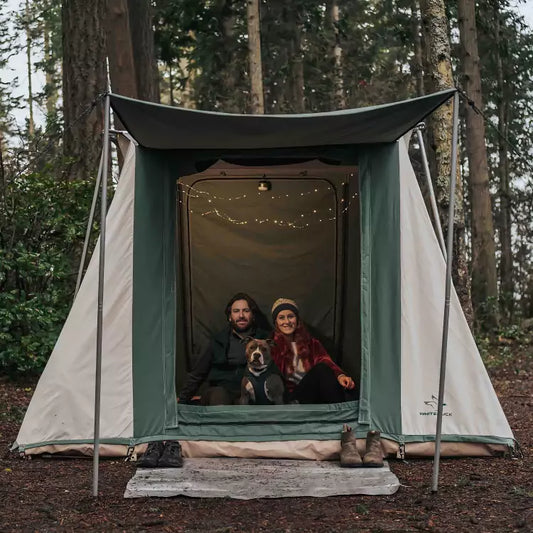 Two people and a dog inside a green and beige tent in a forest setting