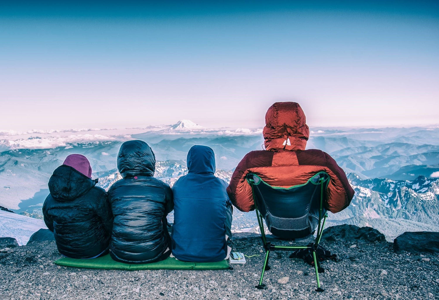 adult sitting in a travel chair on a mountain top with three kids sitting on a mat overlooking mountains with snow on them.
