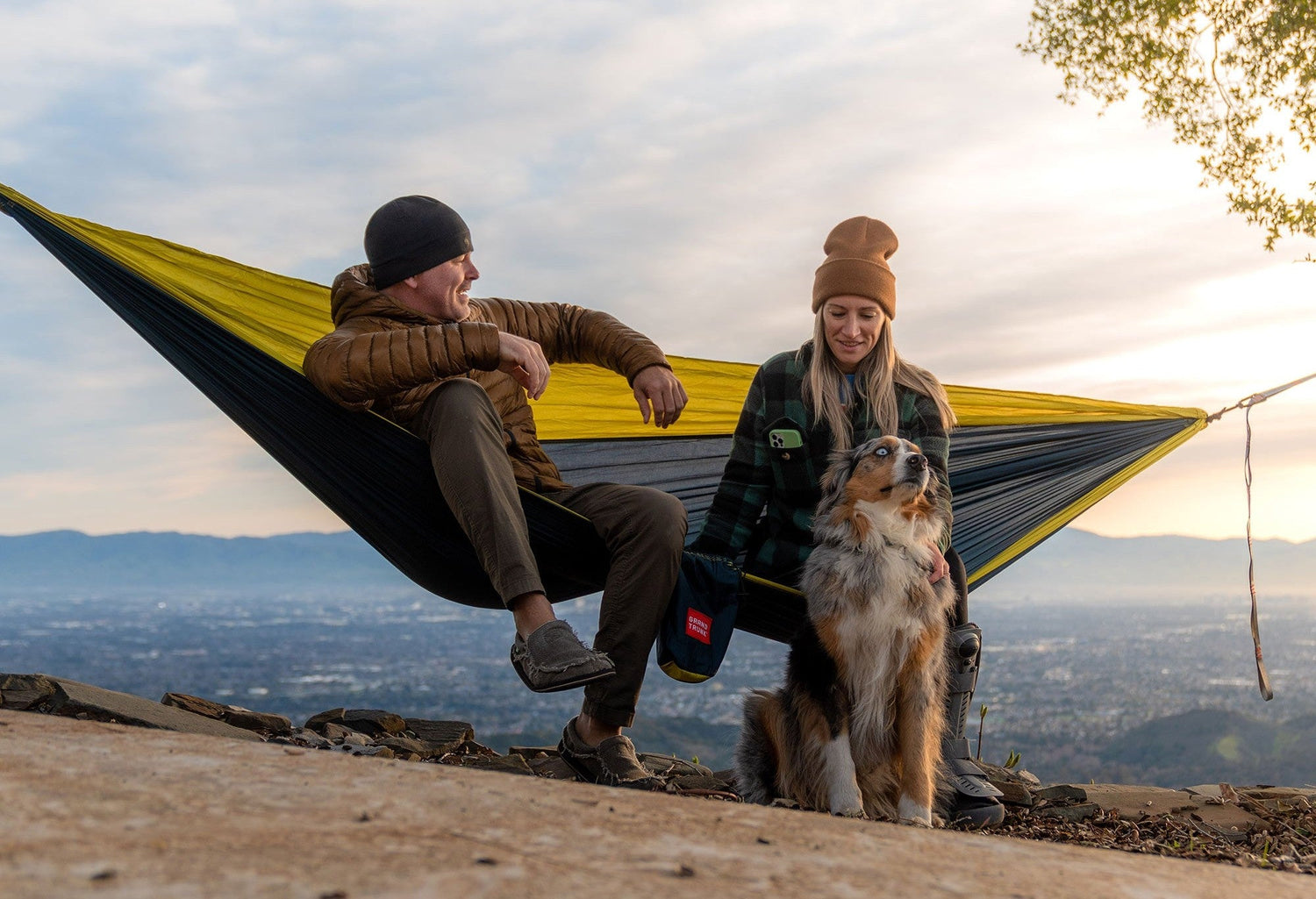 a couple sitting in a Grand Trunk hammock with a dog sitting in front of them. 