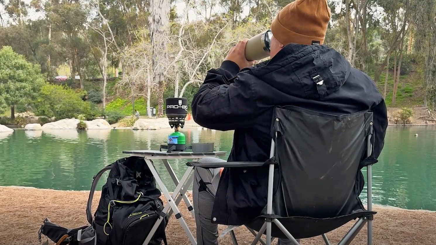 a person sitting in a chair with their back to the camera sipping a cup with a foldable table and backpack in front of them with a lake in the background