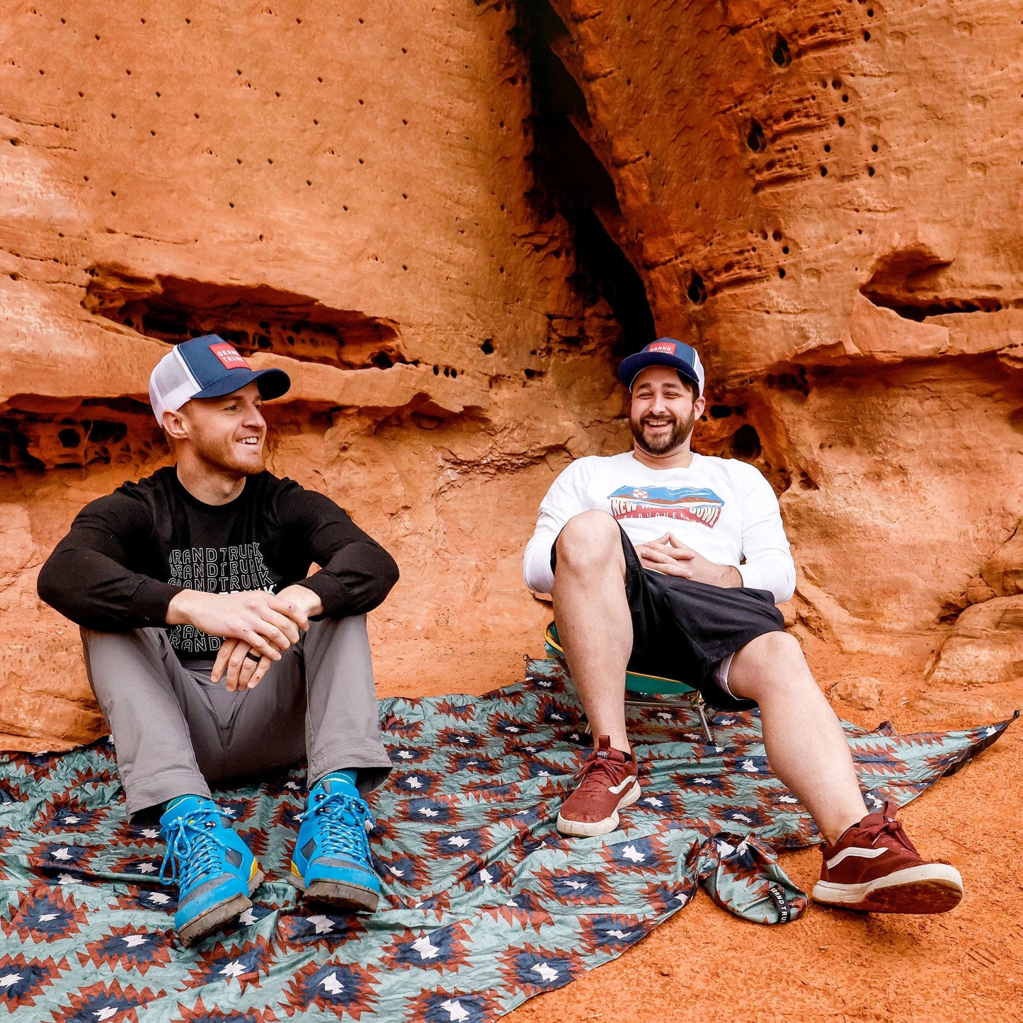 Two men sitting on a patterned blanket against a red rock wall.
