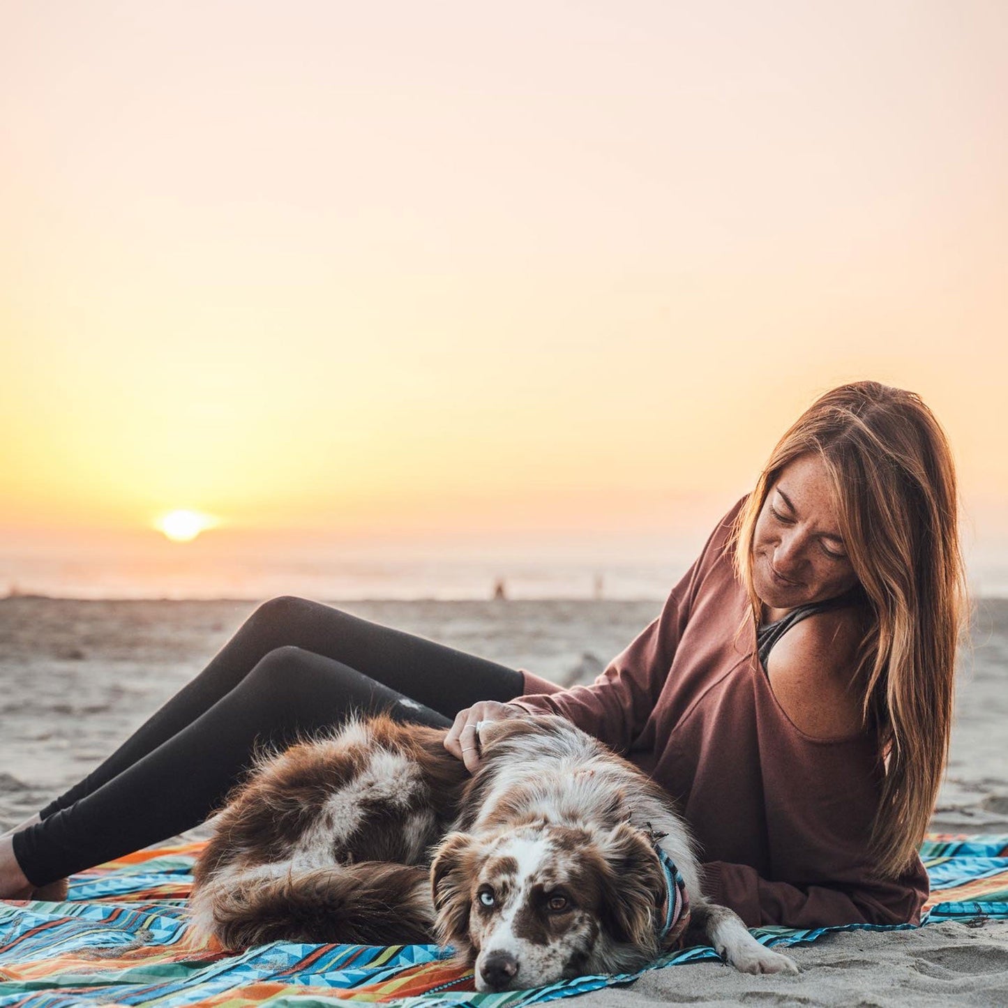 Woman and dog sitting on a beach blanket at sunset