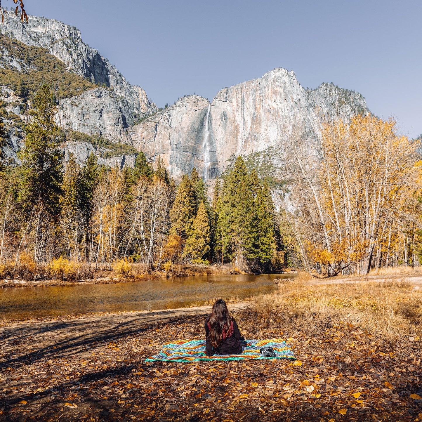 Person sitting on a blanket by a lake with a waterfall and mountains in the background