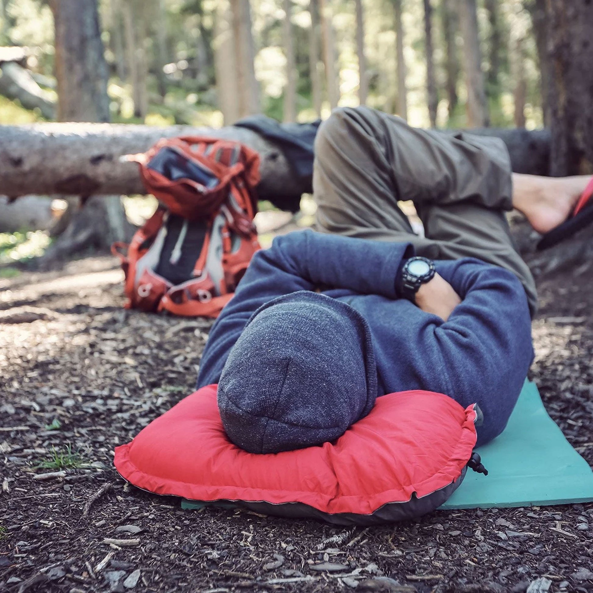 Person lying on a red inflatable pillow in a forest setting with backpack and green mat.