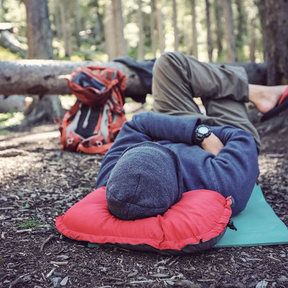 Person lying on a red inflatable pillow in a forest setting with backpack and green mat.