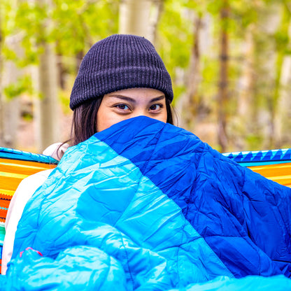 Person wrapped in a blue sleeping bag with a forest background