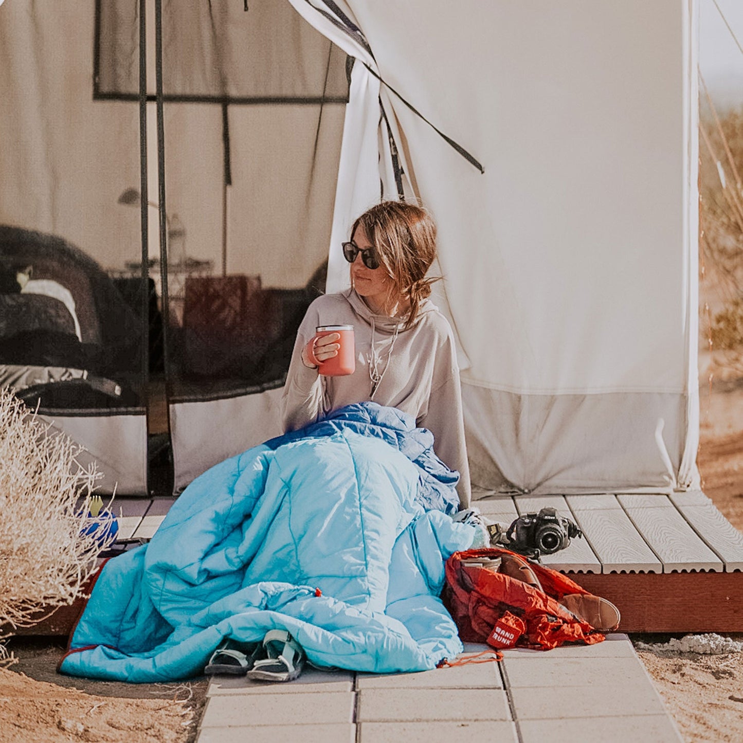 Person sitting in a tent with a blue sleeping bag, holding a drink.