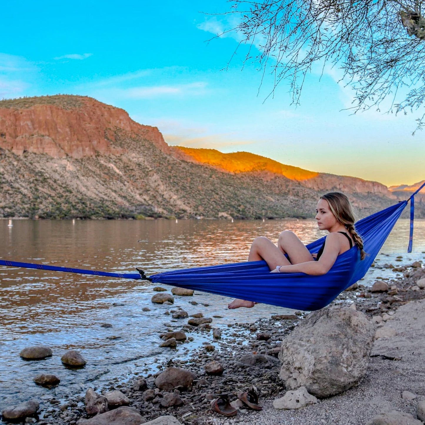 Person sitting in a blue hammock by a lake with mountains in the background