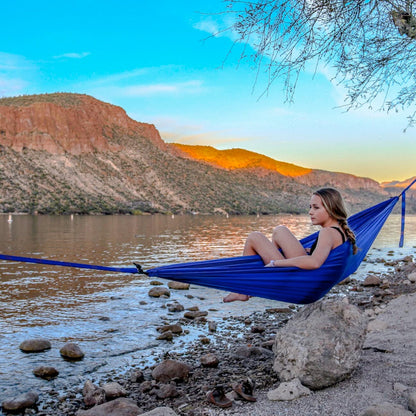 Person sitting in a blue hammock by a lake with mountains in the background