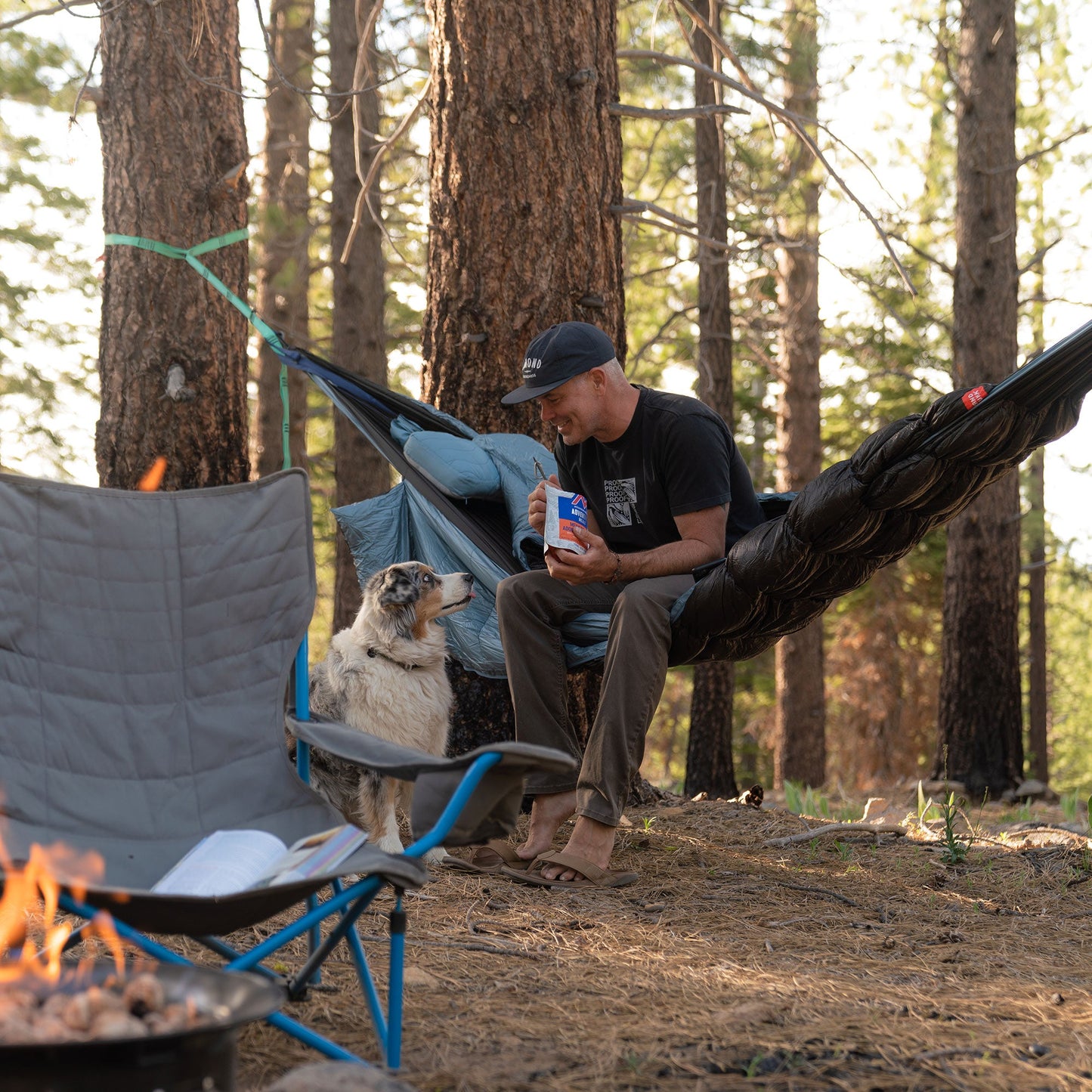 Man sitting in a hammock with a dog by a campfire in a forest setting